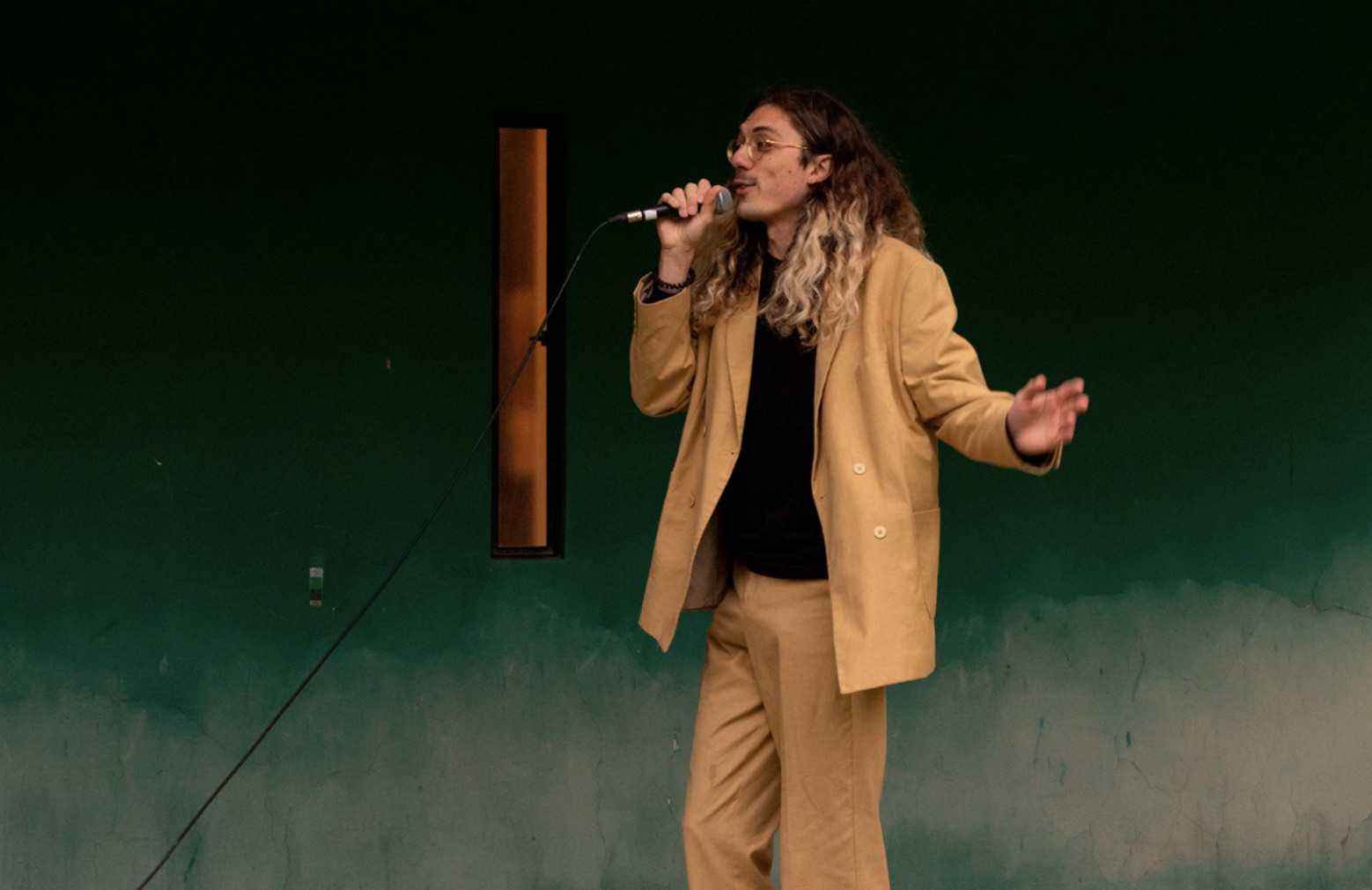 Paul Garcin au micro, en costume beige, devant un mur vert
